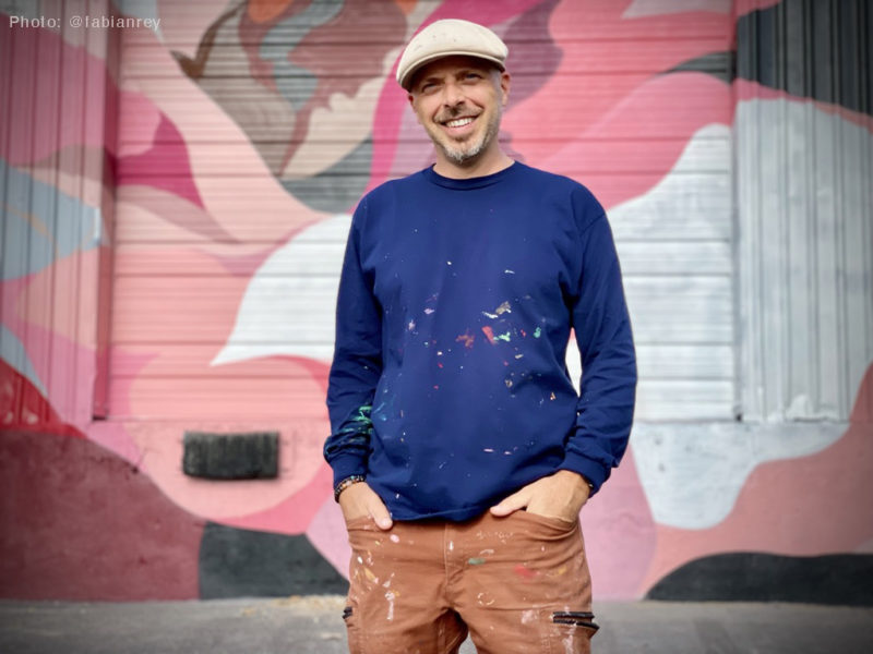 The artist DAAS standing in front of his pink mural outside. He is wearing a tan newsboy hat and has paint splattered on his blue long sleeve shirt