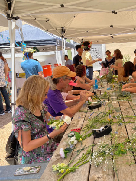 old bakery and emporium's midsummer celebration. photo shows people lined down both sides of a wooden table, putting together flower arrangements