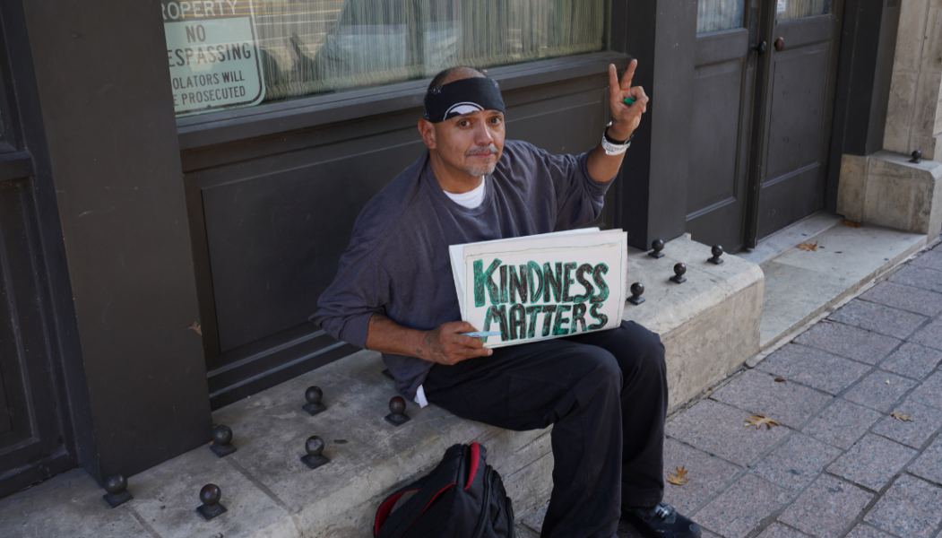 man sitting on 6th street holding a sign that says kindness matters
